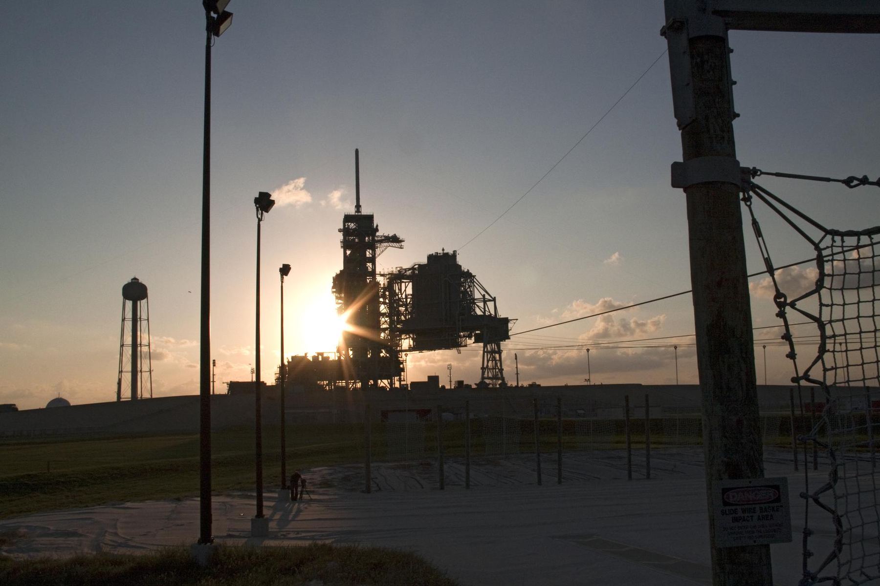 A launch pad tower in silhouette against the rising sun, seen from behind a fence line.