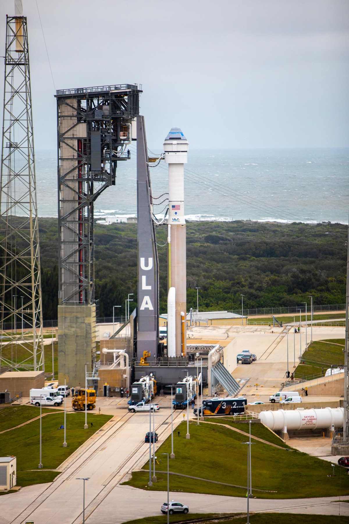 An Atlas V rocket with its crew capsule on top stands beside its service tower on a coastal launch complex.